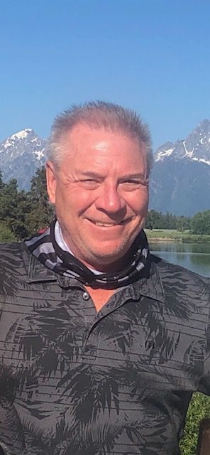 Smiling man outdoors with mountains and lake in background.