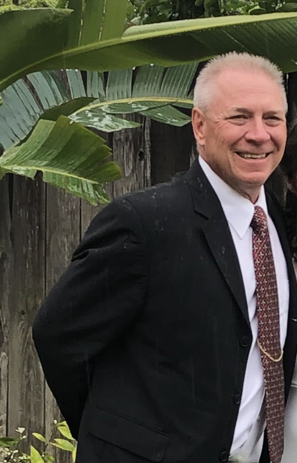 Smiling man in a suit standing outdoors near greenery and a wooden fence.